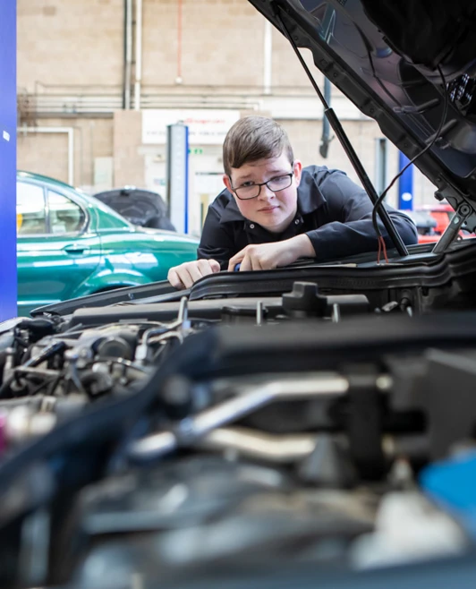 Student in a black overall inspecting a car engine, leaning in closely to examine the mechanical components. Student in a black overall inspecting a car engine, leaning in closely to examine the mechanical components.